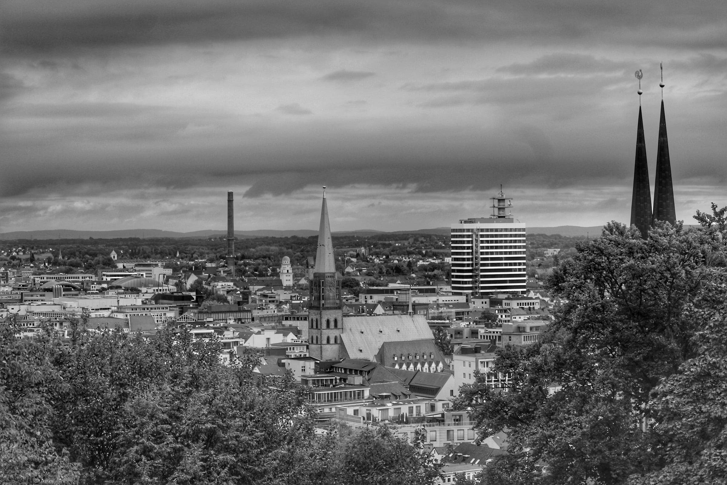 Blick von einer Anhöhe über die Innenstadt von Bielefeld mit Kirchtürmen und Dächern, davor Bäume